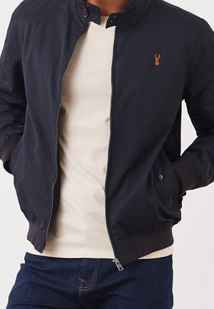 Mann trägt eine marineblaue Jacke mit Reißverschluss und einem beigen Hirsch-Logo auf der Brust, beige Hemd darunter und dunkelblaue Jeans, Hände in den Taschen.