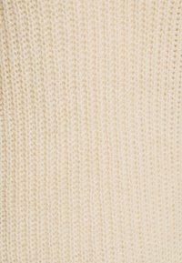 Krämfärgad stickad tyg med en vertikal ribbad struktur, mjuk och tät, som visar en varm nyans och jämn sömnad genom hela ytan.