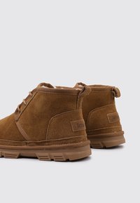 Un par de botas de tobillo de ante marrón con suelas de goma texturizadas y tiradores, mostradas desde la parte posterior sobre un fondo gris claro liso.