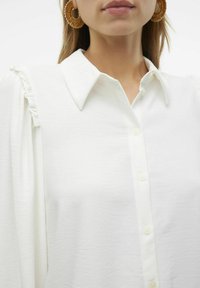 Camicia bianca con colletto, dotata di dettagli arricciati sulle spalle e una texture liscia. Orecchini a cerchio dorati di grandi dimensioni.