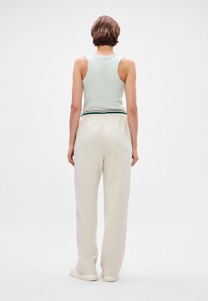 Personne aux cheveux courts et bruns portant un haut sans manches vert clair et un pantalon ample couleur crème, debout de dos sur fond blanc.