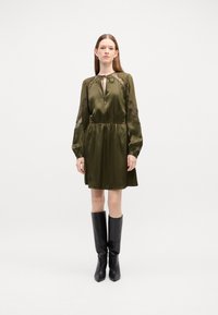 Olijfgroene satijnen jurk met lange mouwen, sleufnek en kanten accenten. Strak bij de taille en een losse rok, gecombineerd met zwarte kniehoge laarzen.
