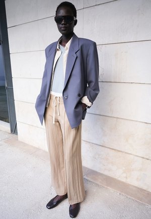 Femeie purtând un blazer supradimensionat lavandă, pantaloni bej cu șnur, top albastru deschis, cămașă în carouri, pantofi negri comozi și ochelari mari de soare închiși la culoare, stând în aer liber.