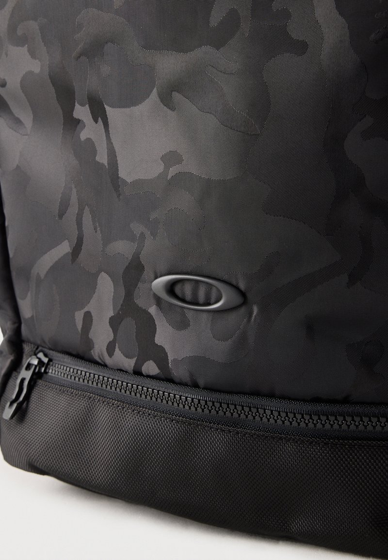 Tessuto nero con motivo mimetico e piccolo logo ovale sopra una cerniera nera su materiale strutturato.