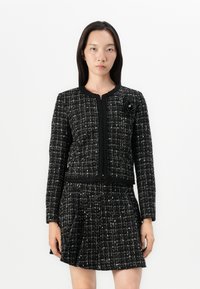 Schwarz-weiße Tweedjacke mit langen Ärmeln, rundem Ausschnitt und strukturiertem Muster. Mit einem floralen Akzent auf der linken Seite.