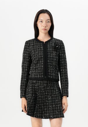 Black and white tweed jacket with long sleeves, a round neckline, and a textured pattern. Features a floral accent on the left side.