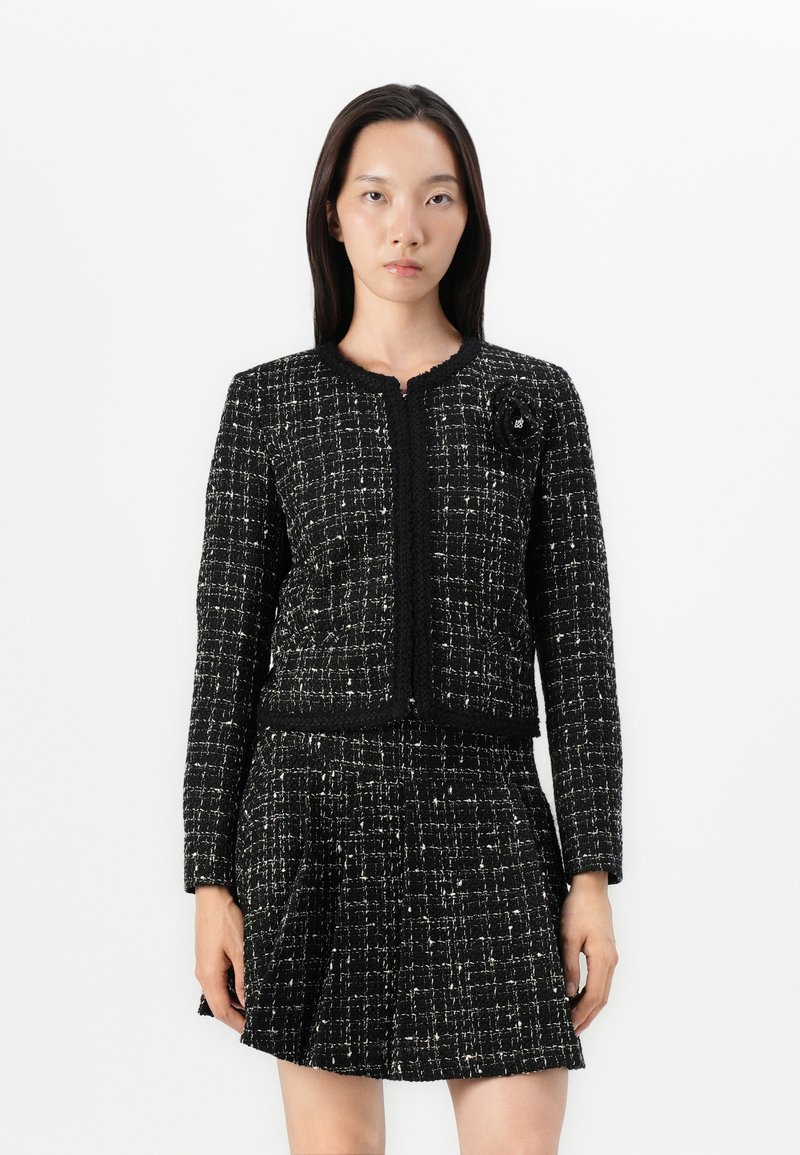 Schwarz-weiße Tweedjacke mit langen Ärmeln, rundem Ausschnitt und strukturiertem Muster. Mit einem floralen Akzent auf der linken Seite.