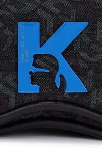 Blå plastbokstav K med en silhuettprofil av en person som bär glasögon på mörk denimtyg med svart läderkant.