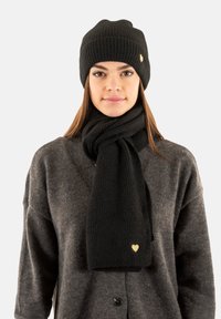 Black knit beanie and scarf set made from soft material. Scarf features a gold heart accent. Model wears a grey cardigan underneath.