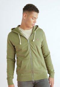 Oliven grønn zip-up hettegenser med snor, frontlommer og ribbet tekstur. Brukt over en lys beige skjorte med et lite logo.
