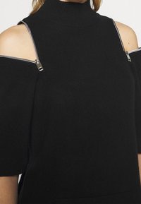 Pull noir en tricot avec col montant, épaules découvertes et fermetures éclair sur les manches. Présente une coupe décontractée et un tissu texturé.