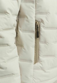 Doudoune beige clair avec des coutures horizontales et une poche zippée. Elle présente une texture lisse et brillante avec de petites perforations.