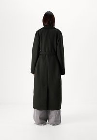 Μακρύ, σκούρο πράσινο παλτό φτιαγμένο από παχύ υλικό, με ζώνη στη μέση και μεγάλες τσέπες. Το παλτό έχει ίσια σιλουέτα και ψηλό γιακά.