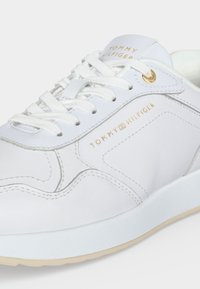 Baskets blanches en cuir Tommy Hilfiger avec lacets blancs, œillets dorés et texte du logo doré sur le côté et la languette.
