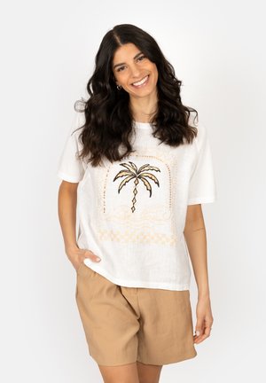 Smiling woman with long dark hair wearing a white T-shirt with a palm tree design and beige shorts, standing with one hand in pocket.