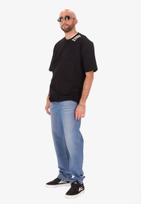 Uomo con barba e orecchini che indossa una t-shirt nera con grafica, jeans blu, sneakers nere e occhiali da sole in piedi contro uno sfondo bianco.