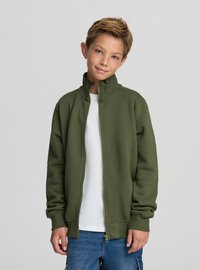 Giacca verde oliva con zip e colletto alto, texture morbida e tasche frontali, indossata sopra una camicia bianca, abbinata a jeans blu.