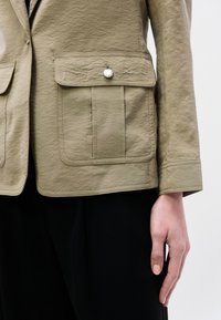 Κοντινό πλάνο ενός ατόμου που φοράει μπεζ σακάκι με τσεπάκι με καπάκι και κουμπί και μαύρο παντελόνι, με το χέρι να ξεκουράζεται δίπλα του.