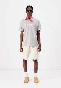 Camiseta gris de manga corta con gráfico de "REPLAY", pantalones cortos beige de cargo, bandana roja y gafas de sol. Unos zapatos marrón claro completan el atuendo.