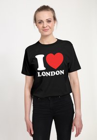 Mladá žena s blond vlasy, oblečená v černém tričku "Miluji Londýn" a černých kalhotách, na jednoduchém bílém pozadí.