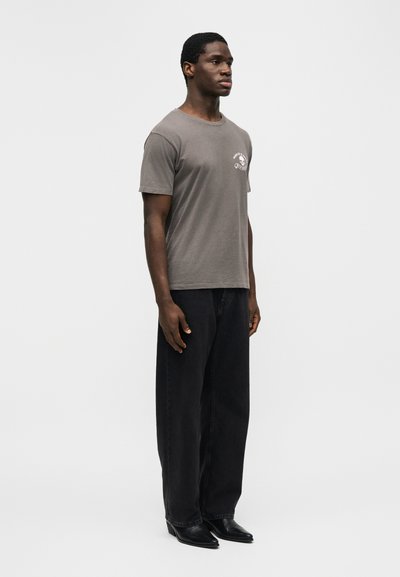 Νέος άνδρας όρθιος, φορώντας γκρι μπλουζάκι με σχέδιο, φαρδιά μαύρη παντελόνια και μαύρα μυτερά παπούτσια, μπροστά σε απλό ανοιχτό φόντο.