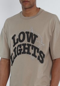 Camiseta de algodón beige con texto negro sobredimensionado que dice "BAJAS LUCES" con un contorno gris, de manga corta y corte relajado.