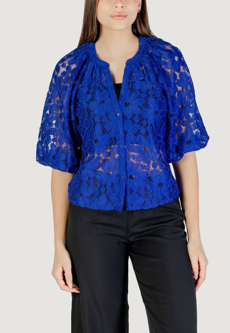 Blusa di pizzo blu con motivi floreali, chiusura a bottoni e maniche a palloncino corte. Abbinata a pantaloni neri, con vestibilità comoda e tessuto liscio.