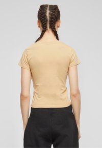 Kortermet, tettsittende t-skjorte i lys beige bomull med rund hals, sett fra baksiden. Glatt tekstur og minimalistisk design.