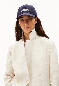 Cappello da baseball blu con testo ricamato bianco "Krawedangels dal 2007." Indossato con una giacca colletto beige chiaro con una texture liscia.