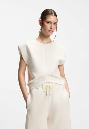 Femme aux cheveux attachés portant un haut blanc sans manches avec épaules rembourrées et un pantalon ample assorti, les mains dans les poches, sur fond blanc.