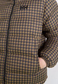 Gros plan sur une main dans la poche d'une doudoune à motifs pied-de-poule marron et noir, avec une fermeture éclair noire et un logo sur la poitrine.