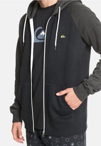 Sudadera con cremallera de color azul marino oscuro, con mangas grises, cordones blancos y dos bolsillos frontales. Presenta un pequeño logo amarillo en el lado izquierdo del pecho.