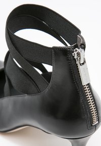 Chaussure à talon en cuir noir avec fermeture éclair argentée à l'arrière et brides croisées élastiques autour de la cheville.