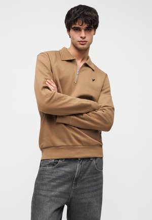 Brązowa bluza rozpinana na zamek z kołnierzem, wykonana z gładkiego materiału, z czarnym logo na piersi i ściągaczami na rękawach, zestawiona z szarymi dżinsami.