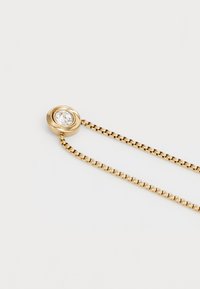 Collar de color dorado con un colgante circular retorcido que cuenta con una gema redonda y transparente. La cadena está compuesta por pequeños segmentos enlazados.