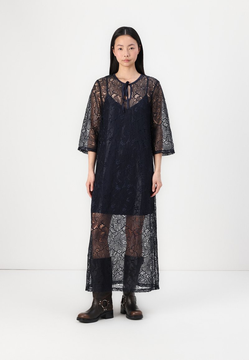 Robe en dentelle bleu marine avec des manches 3/4, texture transparente, motif floral et un détail à nouer au niveau du décolleté, superposée à des collants foncés assortis.