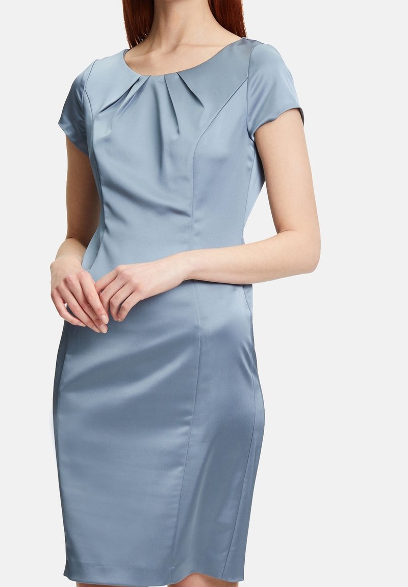Femme portant une robe cintrée en satin bleu clair à manches courtes, avec un col rond et un détail plissé sur la poitrine, mains jointes devant.