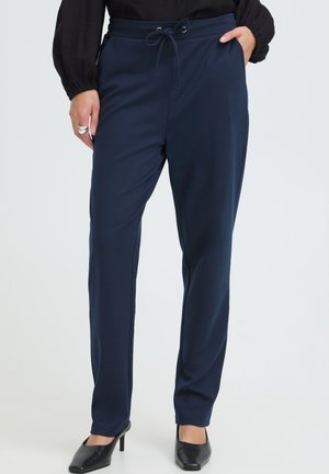 Personne portant un pantalon bleu marine à cordon, un chemisier noir, des talons noirs, une main dans la poche, debout devant un fond clair et uni.