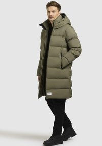 Lang, olivengrøn, polstret frakke med hætte, der har lynlåslommer og et logo-patch. Parret med sorte bukser og støvler.