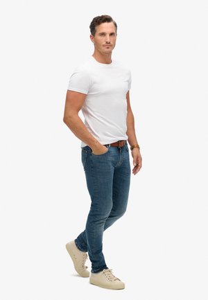 Camiseta blanca de manga corta, pantalones vaqueros azul oscuro, zapatillas de color beige claro. El modelo está de pie con una mano en el bolsillo, mostrando un atuendo casual.