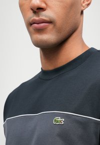 Κοντομάνικο πουκάμισο σε σκούρο μπλε βαμβάκι με υφή, διαθέτοντας μια αντίθετη γκρι πάνελ και κεντημένο πράσινο λογότυπο κροκόδειλου.