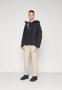 Veste imperméable noire avec doublure grise, poche poitrine zippée, pantalon cargo beige avec zips et chaussures de randonnée noires. Design simple et fonctionnel.