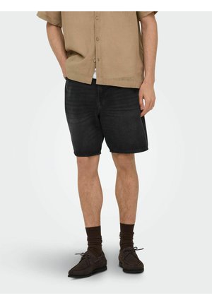 Homme portant un short en jean noir, des chaussettes marron, des chaussures marron foncé lacées et une chemise beige à manches courtes boutonnée, debout devant un fond uni.