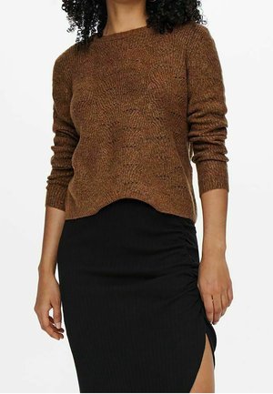 Femme portant un pull en maille marron à manches longues et une jupe noire avec une fente froncée sur le côté, visible des épaules jusqu'au milieu des cuisses.