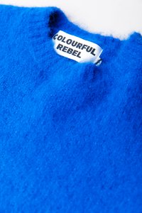 Taflă albastră strălucitoare și pufoasă, cu o etichetă albă de îmbrăcăminte care scrie "COLOURFUL REBEL" cu litere mari negre.