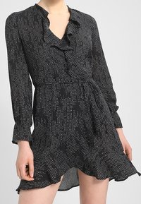 Vestido negro envolvente con un patrón de puntos rectangulares blancos, cuello y dobladillo con volantes, mangas largas y cinturon en la cintura, luciendo una persona de pie.