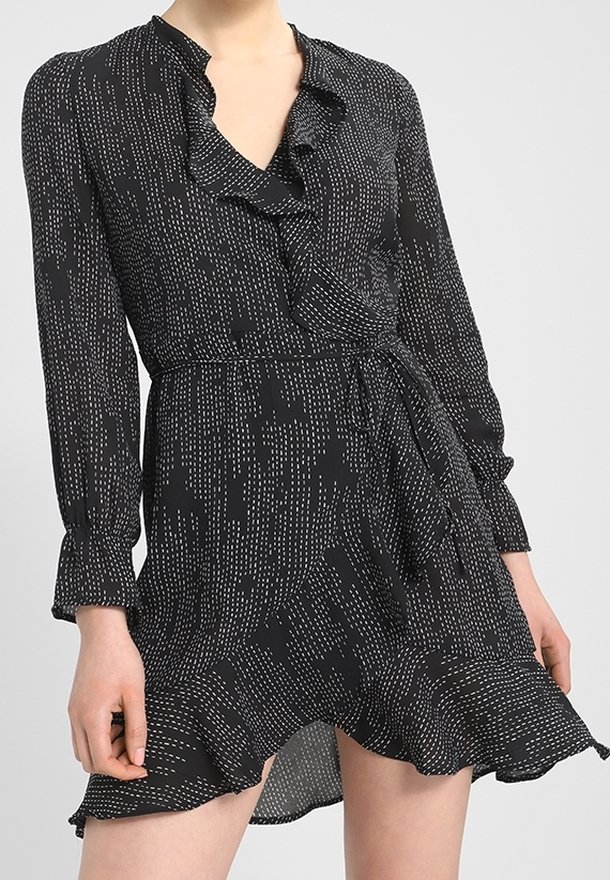 Vestido negro envolvente con un patrón de puntos rectangulares blancos, cuello y dobladillo con volantes, mangas largas y cinturon en la cintura, luciendo una persona de pie.