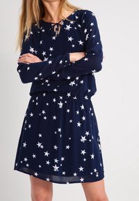 Kvinna klädd i en marinblå långärmad klänning med vita stjärnmönster, med armarna i kors, framför en enkel ljus bakgrund.