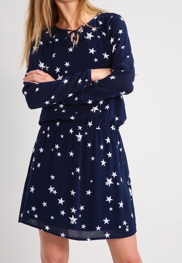 Kvinna klädd i en marinblå långärmad klänning med vita stjärnmönster, med armarna i kors, framför en enkel ljus bakgrund.