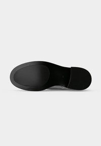 Semelle de chaussure noire avec des zones de prise texturées au talon et à la plante du pied, présentée à plat sur un fond gris clair.